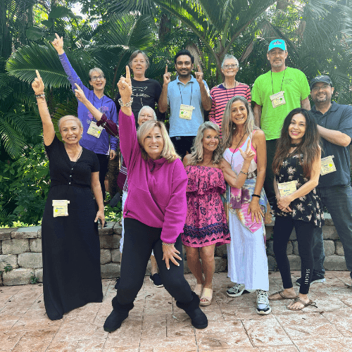 Group of people posing outdoors with hands raised, smiling in a tropical setting.