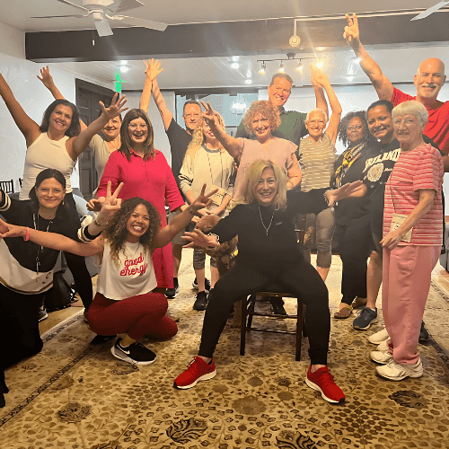 Group of smiling people posing energetically with arms raised in a cheerful indoor setting.